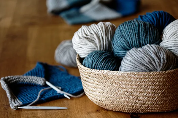 Ein Korb mit mehreren Wollknäueln in Weiß-, Grau- und Blautönen steht auf einem Holztisch. Daneben liegen zwei begonnene Strickstücke in Blau und Grau sowie eine Stricknadel. Im Hintergrund sind weitere Wollknäuel unscharf sichtbar.Bild verlinkt zur Seite