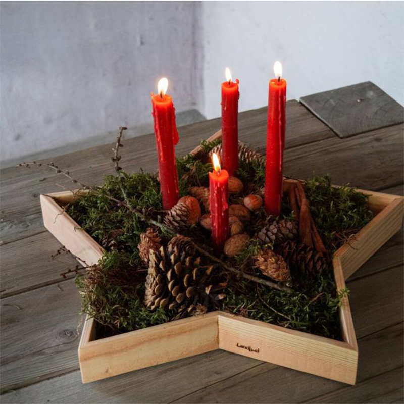 Ein Holztablett in Form eines fünfzackigen Sternes ist mit vier roten Kerzen, Tannengrün und Nüssen dekoriert.