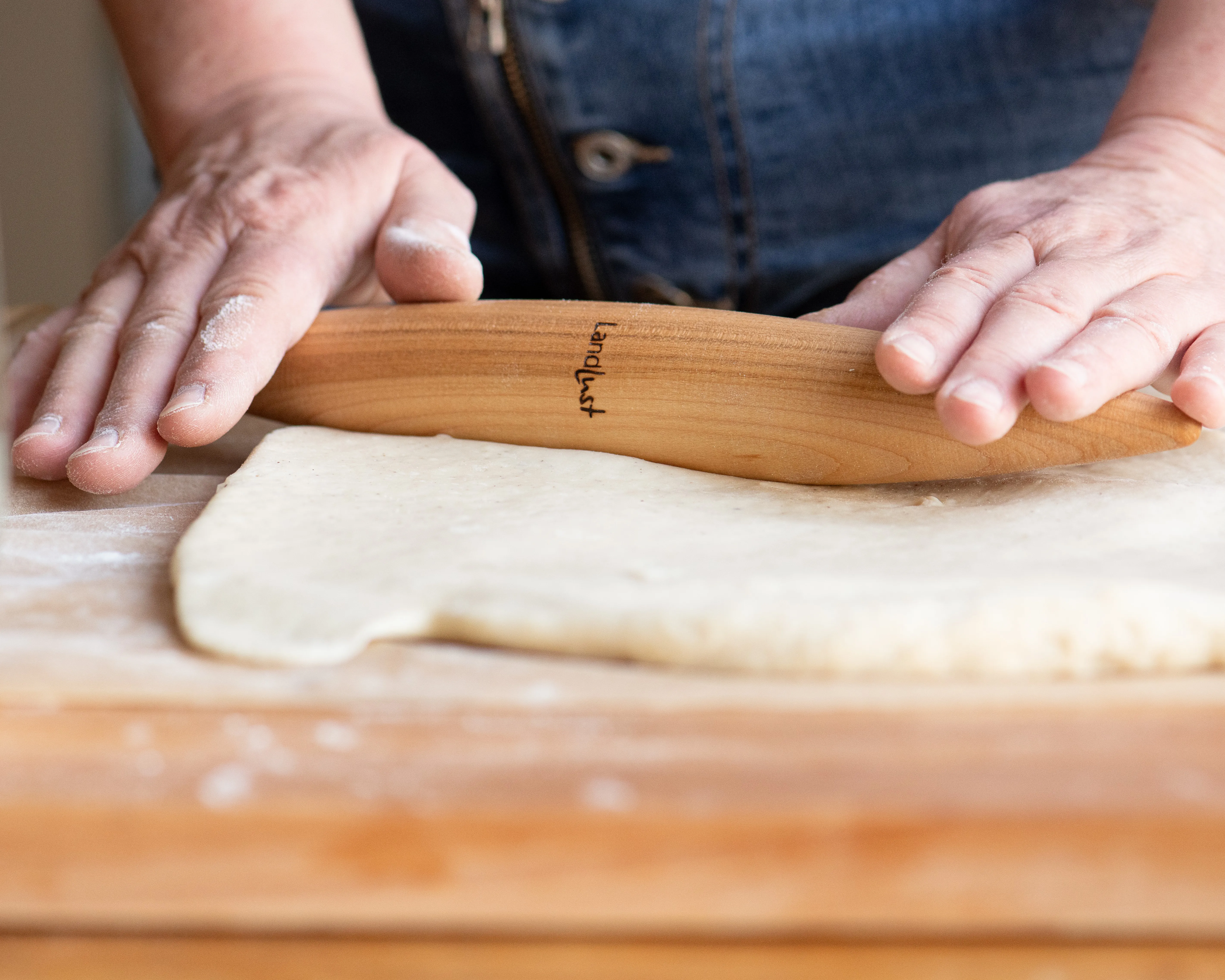 Hände rollen mit einem hölzernen Teigroller Teig auf einem Holzbrett aus. Der Teigroller trägt eine Aufschrift. Die Person trägt dunkle Kleidung und der Fokus liegt auf der Handlung des Teigausrollens. Bild verlinkt zur Seite Küche.