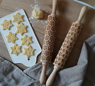 Zwei Teigrollen mit denen die Motive Stern und Schneeflocke auf Kekse übertragen werden können. Auf der bemehlten Holztischplatte liegen auch Keksrolinge, eine Teigkugel und eine graue Tischdecke. 