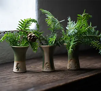 Drei kleine grüne Vasen mit Prägung in Tannenbaum-Form stehen auf einer Fensterbank. Sie sind mit grünen Tannenzweigen dekoriert.