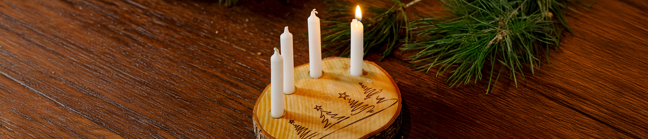 Vier Kerzen sind auf eine Holzscheibe gesteckt, in die Tannenbäume geritzt sind. Das ganze steht auf einem Holztisch auf dem auch Tannenzweige und goldene Weihnachtskugeln liegen
