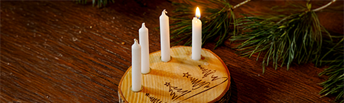 Vier Kerzen sind auf eine Holzscheibe gesteckt, in die Tannenbäume geritzt sind. Das ganze steht auf einem Holztisch auf dem auch Tannenzweige und goldene Weihnachtskugeln liegen