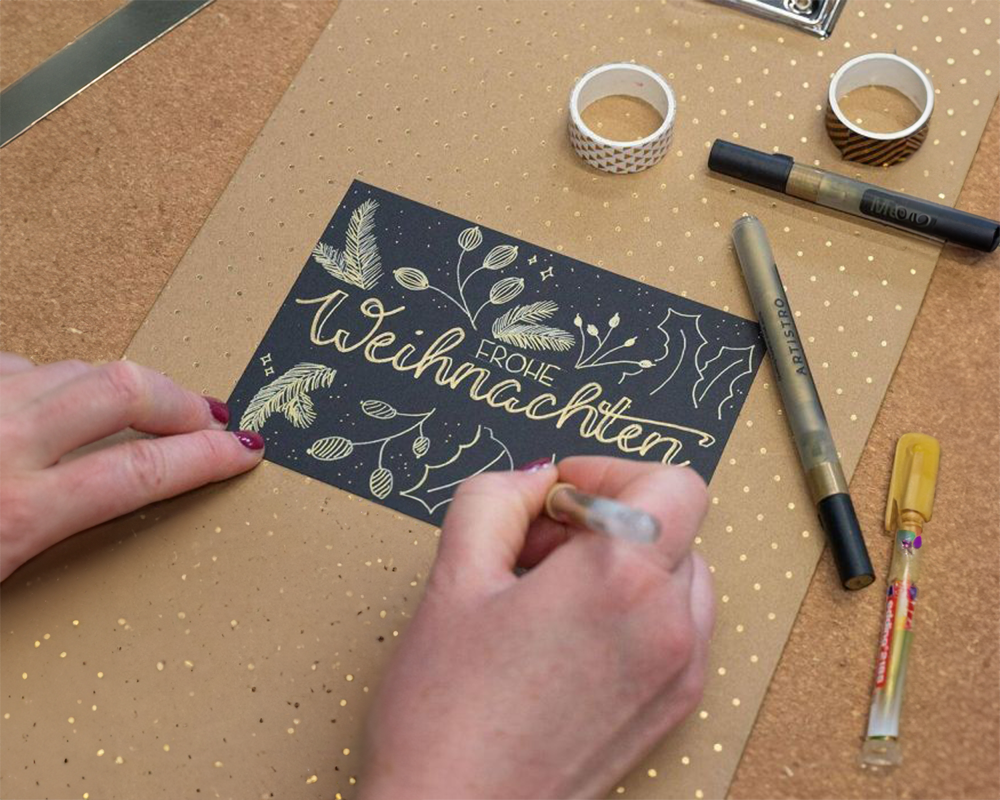 Eine Hand schreibt auf einer schwarzen Karte mit weihnachtlichem Design. Im Hintergrund ist braunes Papier und verschiedene Stifte sowie andere Bastelutensilien