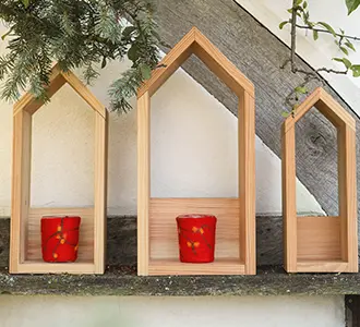 Drei offene Holz-Windlichter in Form von Häusern in unterschiedlichen Größen stehen vor einem Fachwerkhaus. Im Inneren der Windlichter befinden sich rote Kerzengläser.