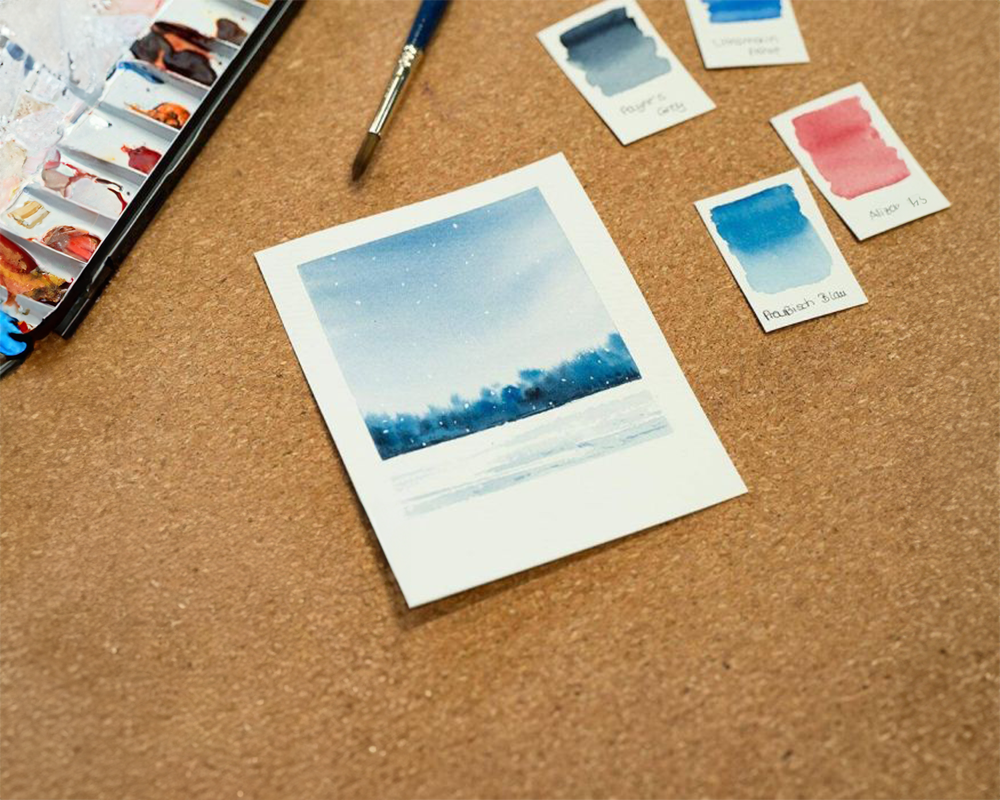 Auf einem kleinen Aquarellblatt ist eine blau-weiße Winterlandschaft zu sehen. Das Blatt liegt auf einer braunen Unterlage. Daneben ein Farbkasten sowie Farbteststreifen.