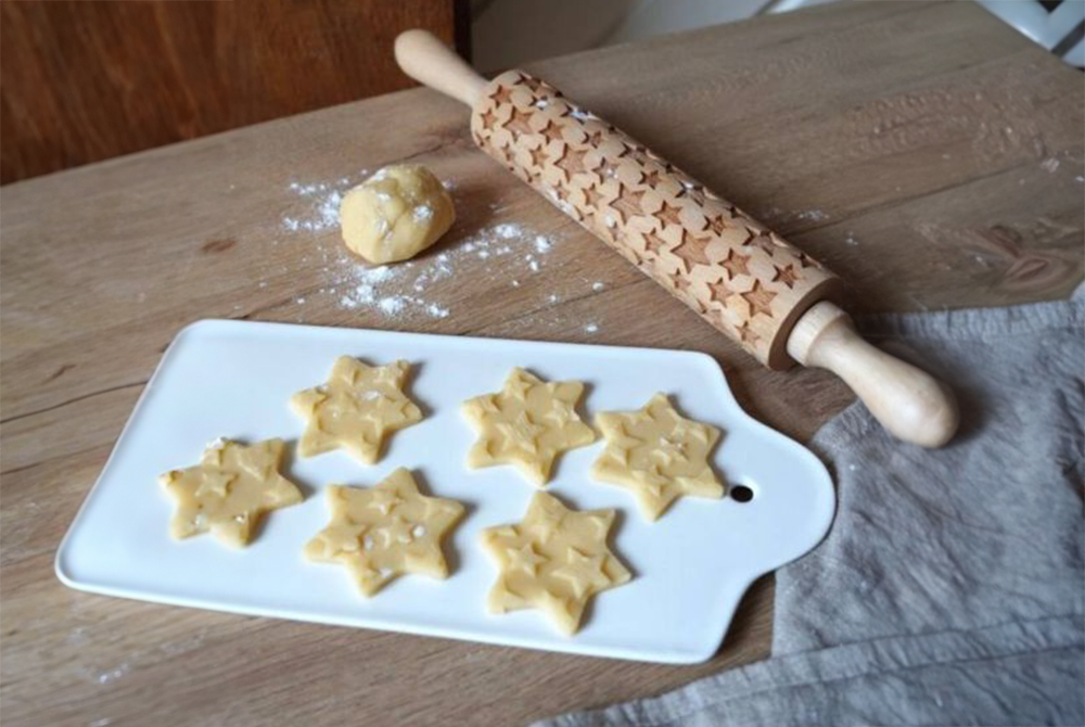 Eine Teigrolle mit Stern-Motiv liegt neben einem Brett mit rohen, ausgestochenen Plätzchen und einem grauen Tuch auf einem Holztisch