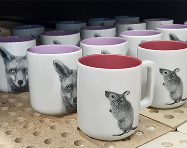 Tassen mit unterschiedlichen Tiermotiven und Farben stehen nebeneinander in einem Brennofen.