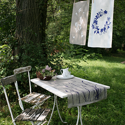 Auf einem gedeckten Tisch liegt ein bedrucktes Tischtuch. Dahinter vom Baum hängen weitere Tücher mit anderen Motiven.