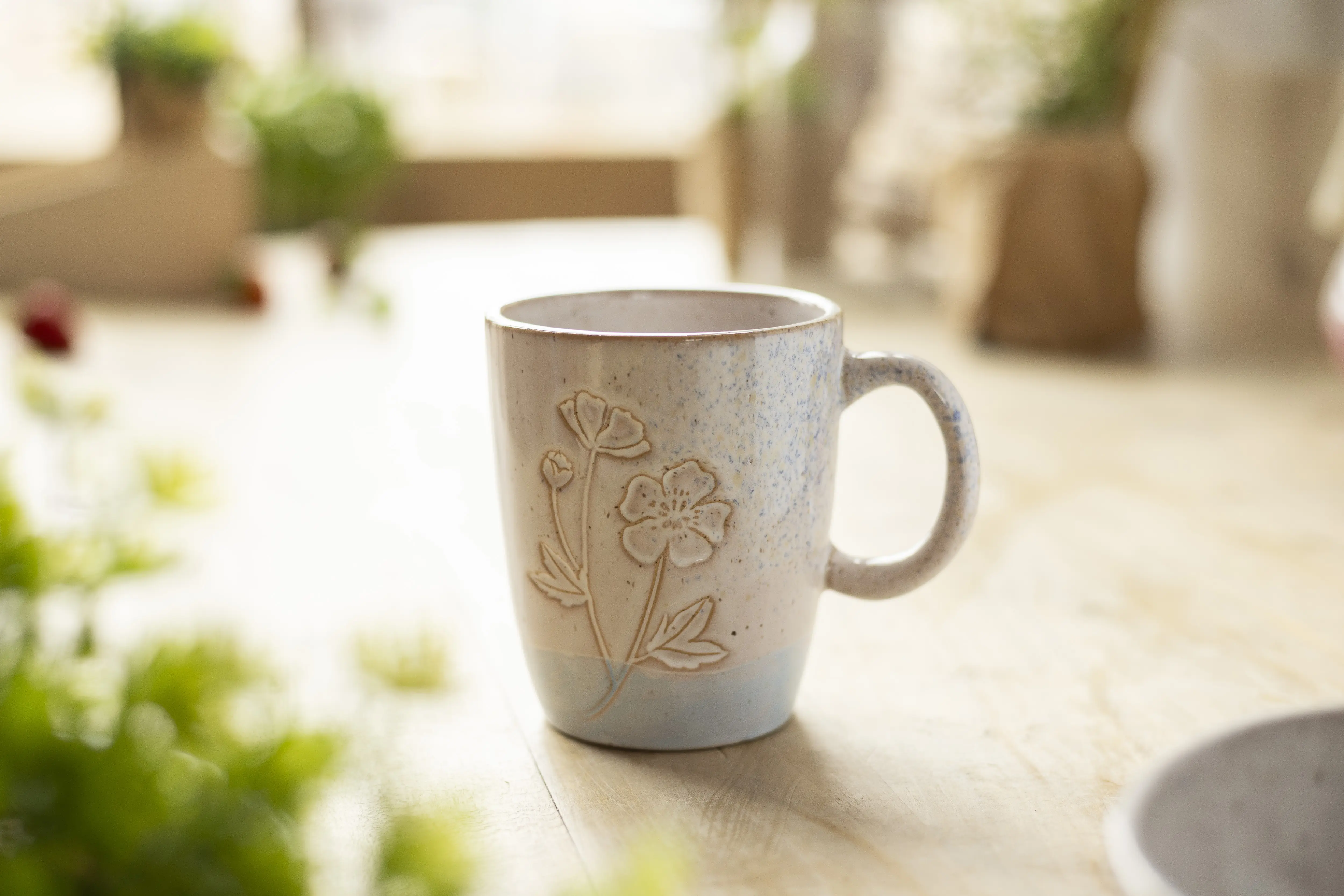 Eine beige Keramiktasse auf einem mit Pflanzen dekorierten Tisch, die ab dem letzten unteren Drittel Pastellblau angemalt ist. Verziert ist die Tasse mit einem Blumenmotiv. 