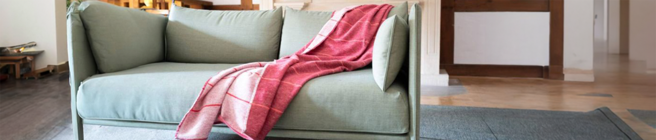 Zwei Personen sitzen in beigen Pullovern auf einem blauen Sofa, zugedeckt mit einer gemusterten Decke, halten je eine Tasse. Neben ihnen liegt ein aufgeschlagenes Buch. Im Hintergrund steht eine Topfpflanze.