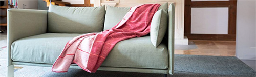 Zwei Personen sitzen in beigen Pullovern auf einem blauen Sofa, zugedeckt mit einer gemusterten Decke, halten je eine Tasse. Neben ihnen liegt ein aufgeschlagenes Buch. Im Hintergrund steht eine Topfpflanze.