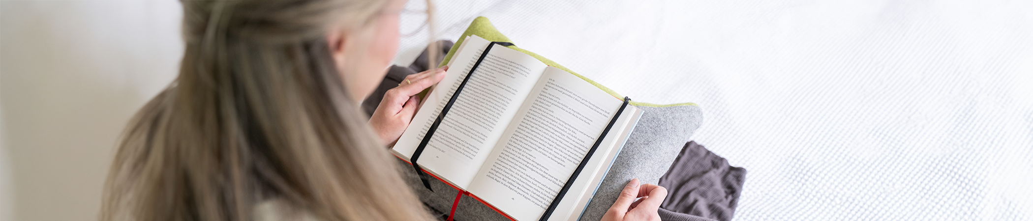Person sitzt und liest ein Buch. Das Buch ist auf einem speziellen Lesekissen, dass auf dem Schoß der Person liegt, befestigt.