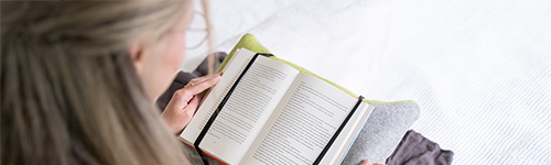 Person sitzt und liest ein Buch. Das Buch ist auf einem speziellen Lesekissen, dass auf dem Schoß der Person liegt, befestigt.