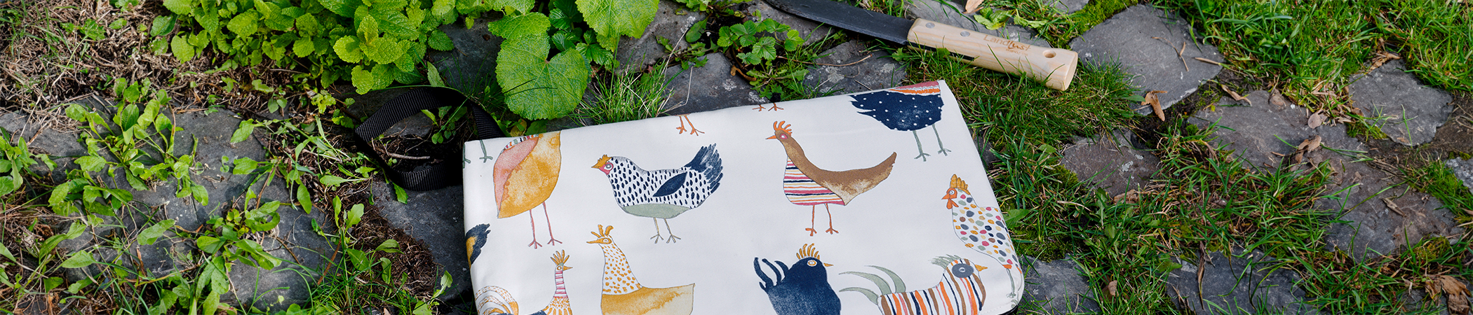 Ein Kniekissen mit Hühneraufdruck zum Gärtnern liegt neben anderen Gartenwerkzeugen auf einem bewachsenen Boden