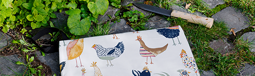 Ein Kniekissen mit Hühneraufdruck zum Gärtnern liegt neben anderen Gartenwerkzeugen auf einem bewachsenen Boden