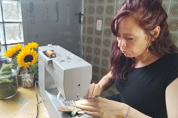 Eine Frau mit langen braunen Haaren näht an einer Nähmaschine auf einem Tisch. Neben ihr steht eine Vase mit Sonnenblumen. Im Hintergrund sind eine graue Wand und Fliesen zu sehen. Bild verlinkt zum Herstellerportrait. 