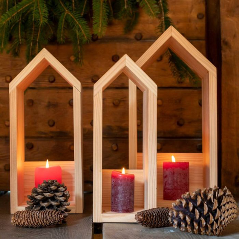 Drei offene Holz-Windlichter in Form von Häusern in unterschiedlichen Größen stehen vor einem Fachwerkhaus. Im Inneren der Windlichter befinden sich rote Kerzengläser.