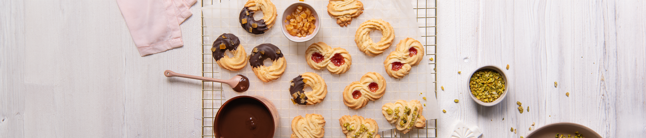 Auf einem hellen Holztisch steht ein Backrost. Darauf liegt verschiedenes Spritzgebäck, dass mit gehackter Pistazie und geschmolzener Schokolade verziert wird
