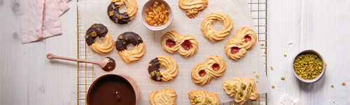 Auf einem hellen Holztisch steht ein Backrost. Darauf liegt verschiedenes Spritzgebäck, dass mit gehackter Pistazie und geschmolzener Schokolade verziert wird