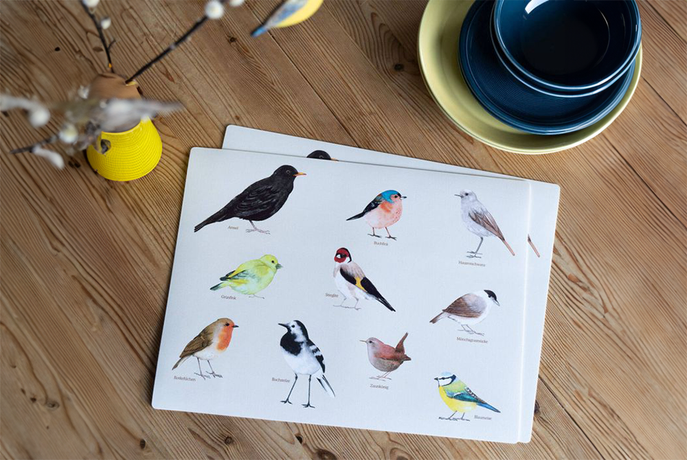 Auf einem Holztisch liegen zwei weiße Tischsets mit Vogel-Print. Daneben eine gelbe Vase und etwas Geschirr
