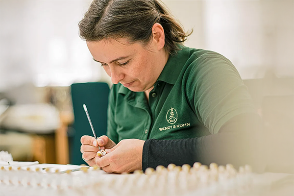Eine Person in grüner Arbeitskleidung bemalt sorgfältig ein kleines Objekt mit einem feinen Pinsel. Im Vordergrund sind unscharfe Objekte zu sehen. Bild verlinkt zur Herstellerseite.
