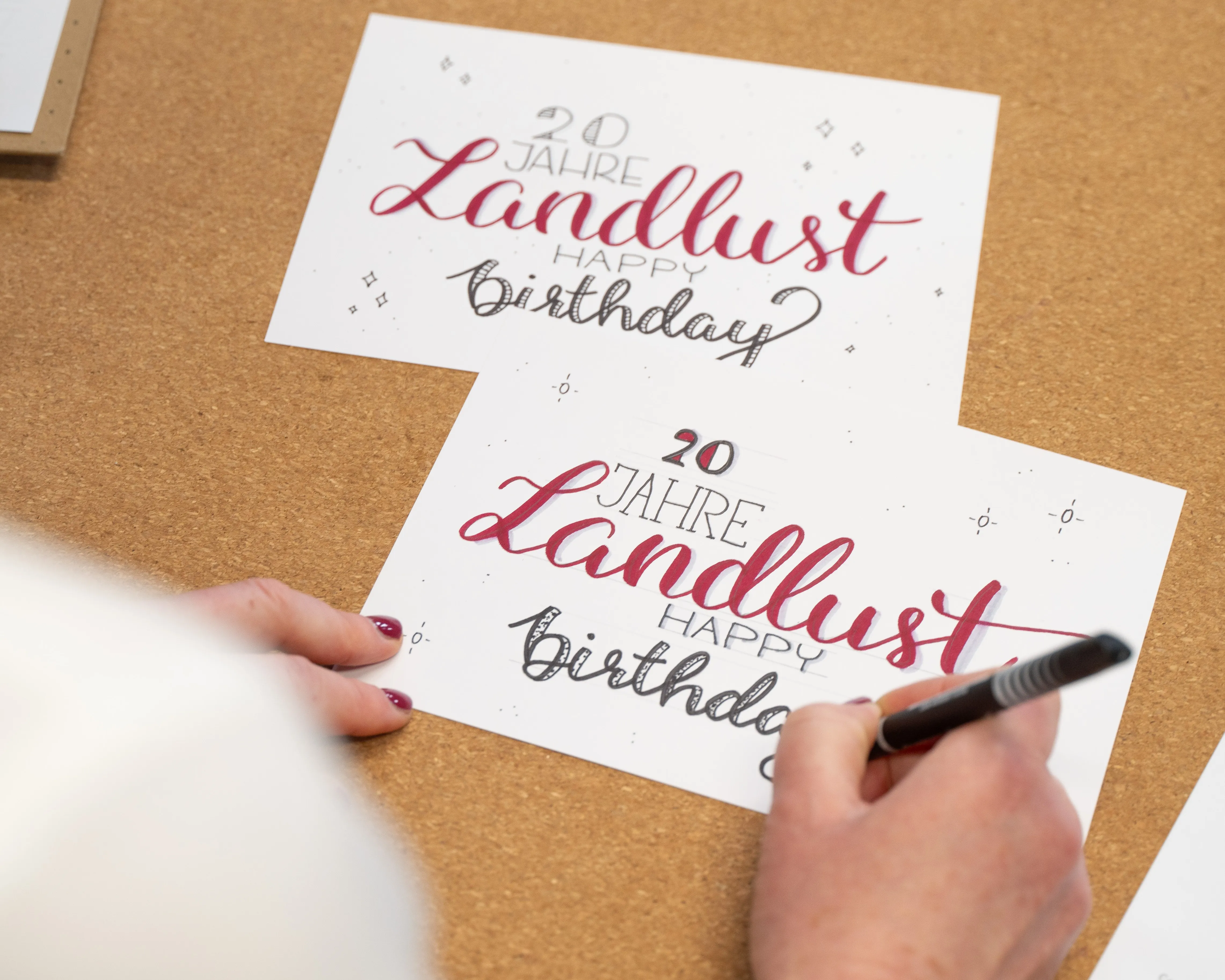 Eine Hand hält einen Stift und schreibt auf eine Karte mit der Aufschrift "20 Jahre Landlust - Happy Birthday" in roter und schwarzer Schrift. Im Hintergrund befindet sich eine identische Karte, Bild verlinkt zum Workshop.