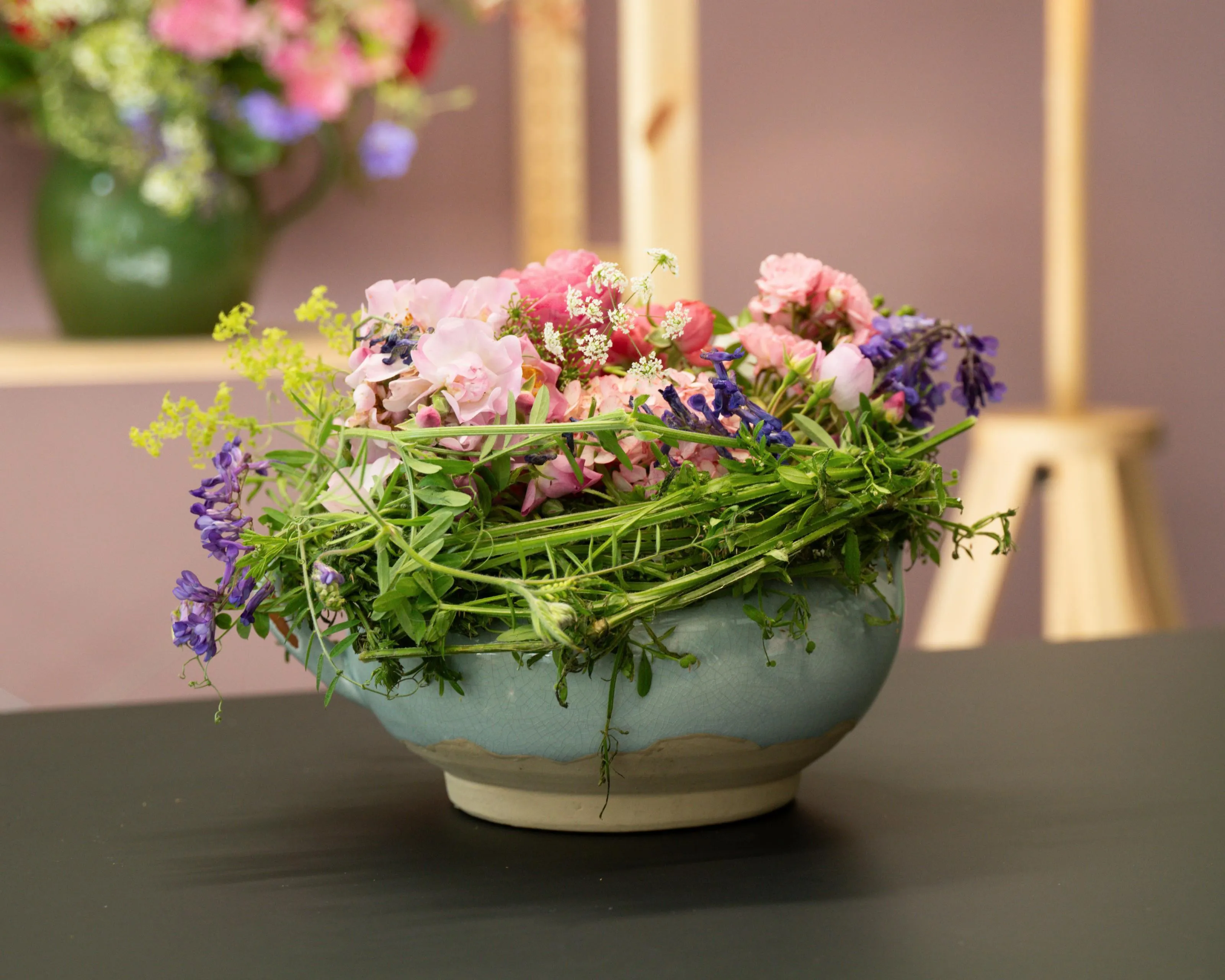Blumenarrangement in einem Nest aus Zweigen mit violetten und rosa Blüten sowie grünen Blättern, dekoriert mit einer Feder auf rosa Hintergrund, Bild verlinkt zum Workshop.
